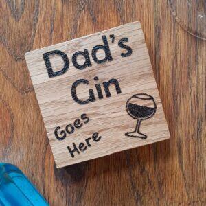 The image shows a oak coaster with the words 'Dad's Gin Goes Here' hand burnt on it. The coaster has a hand burnt gin glass.
