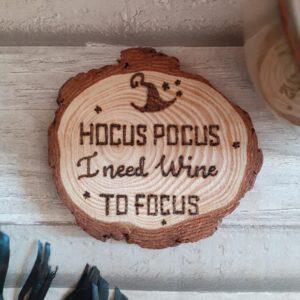 A circular wood slice coaster featuring hand-burnt text that reads 'Hocus Pocus I Need Wine to Focus.' The coaster showcases a natural wood grain pattern, with a rich brown color and a smooth, polished surface. The text is carefully engraved with intricate detail, adding a touch of whimsy to the design. The coaster serves as a functional and decorative piece, perfect for holding a glass of wine while adding a playful and humorous element to any setting."