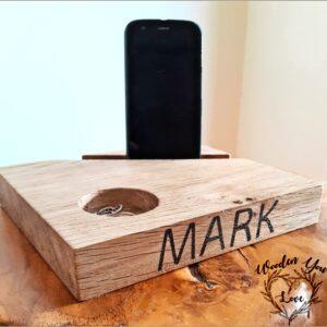 A solid oak mobile phone stand for a bedside table. The stand is crafted from high-quality oak wood, showcasing its natural grain patterns and warm brown hue. Designed to securely hold a smartphone, the stand features a sleek and minimalist design, with a slightly angled surface that provides a comfortable viewing angle. Its sturdy construction ensures stability, while the smooth finish adds a touch of elegance. This stand is a practical and stylish addition to any bedside table, keeping your phone easily accessible and enhancing the overall aesthetic of your space."