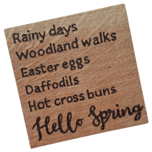 A square wooden coaster featuring intricate oak woodwork with the words 'Hello Spring' engraved on the surface in elegant script. The coaster is handcrafted with attention to detail, showcasing the warmth and natural beauty of wood grain
