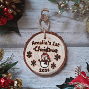 A personalized rustic Christmas tree ornament with a hand burnt snowman design, celebrating a baby's 1st Christmas in 2024. The ornament features a wooden round shape with a charmingly weathered appearance. At the center, a meticulously handcrafted snowman is etched onto the wood, showcasing a cheerful face and a carrot-shaped nose. The snowman wears a cozy hat and scarf, adding a touch of warmth to the design. This sentimental keepsake is a perfect way to commemorate a little one's first holiday season