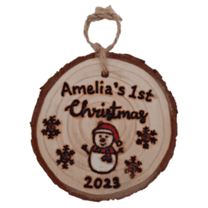 A personalized rustic Christmas tree ornament with a hand burnt snowman design, celebrating a baby's 1st Christmas in 2023. The ornament features a wooden round shape with a charmingly weathered appearance. At the center, a meticulously handcrafted snowman is etched onto the wood, showcasing a cheerful face and a carrot-shaped nose. The snowman wears a cozy hat and scarf, adding a touch of warmth to the design. This sentimental keepsake is a perfect way to commemorate a little one's first holiday season