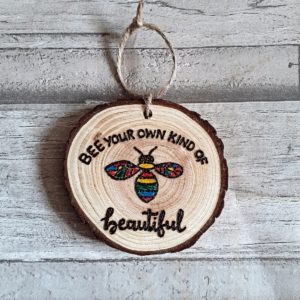 Handcrafted wood slice featuring a rainbow-colored bee design and the inscription "Bee your own beautiful."