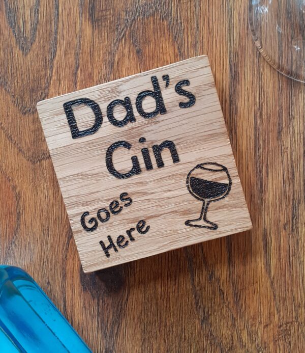The image shows a oak coaster with the words 'Dad's Gin Goes Here' hand burnt on it. The coaster has a hand burnt gin glass.