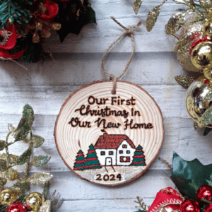 A charming rustic Christmas tree ornament featuring a festive house design. The ornament is shaped like a cozy home with a chimney, adorned with festive decorations. The text on the ornament reads, "Our First Christmas in Our New Home 2024," signifying a special celebration of a couple or family commemorating their first Christmas in their new house.