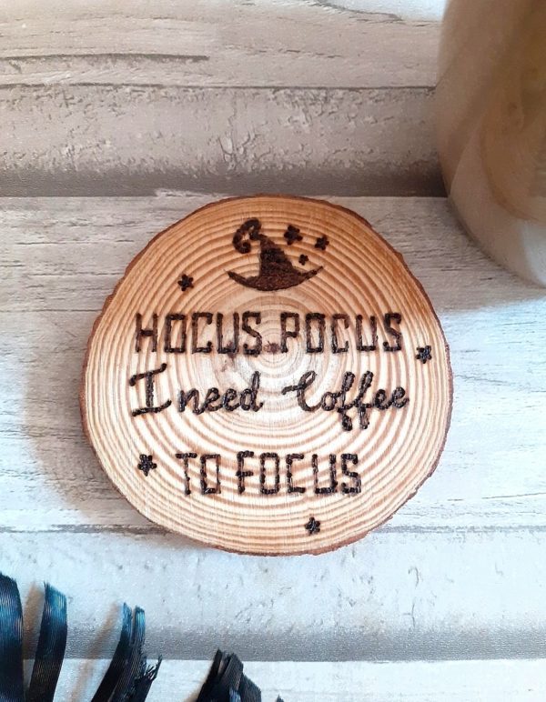 A wooden coaster with a circular shape, featuring a hand-burnt text that reads, "Hocus Pocus I need coffee to focus." The coaster has a natural wood grain pattern, and the burnt text stands out against the background.