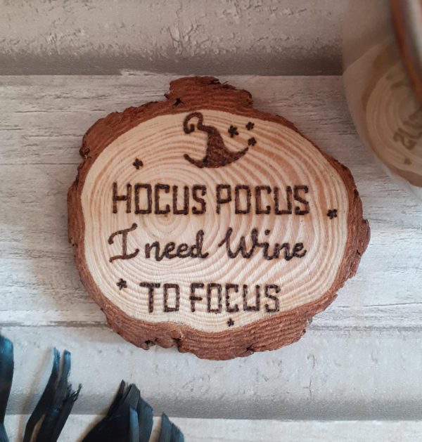 A circular wood slice coaster featuring hand-burnt text that reads 'Hocus Pocus I Need Wine to Focus.' The coaster showcases a natural wood grain pattern, with a rich brown color and a smooth, polished surface. The text is carefully engraved with intricate detail, adding a touch of whimsy to the design. The coaster serves as a functional and decorative piece, perfect for holding a glass of wine while adding a playful and humorous element to any setting."
