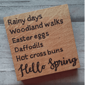 A square wooden coaster featuring intricate oak woodwork with the words 'Hello Spring' engraved on the surface in elegant script. The coaster is handcrafted with attention to detail, showcasing the warmth and natural beauty of wood grain