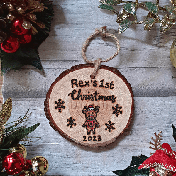 A personalized rustic Christmas tree ornament for a baby's 1st Christmas in 2023. The ornament features a hand-burnt reindeer design on a wooden surface. The reindeer is intricately crafted, showcasing antlers, a gentle face, and a joyful expression. The rustic charm is enhanced by the wood's natural grain and texture. The ornament is delicately engraved with the baby's name and '1st Christmas' to commemorate this special occasion. It hangs gracefully from jute rope, ready to add warmth and merriment to any Christmas tree