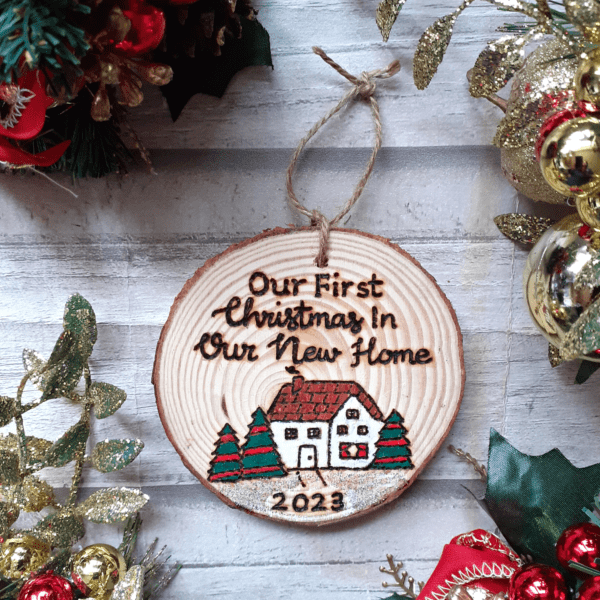 A charming rustic Christmas tree ornament featuring a festive house design. The ornament is shaped like a cozy home with a chimney, adorned with festive decorations. The text on the ornament reads, "Our First Christmas in Our New Home 2023," signifying a special celebration of a couple or family commemorating their first Christmas in their new house.