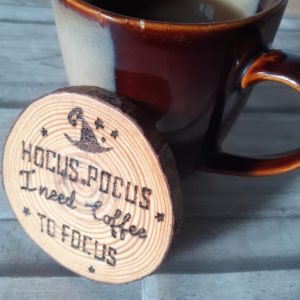 A wooden coaster with a circular shape, featuring a hand-burnt text that reads, "Hocus Pocus I need coffee to focus." The coaster has a natural wood grain pattern, and the burnt text stands out against the background.