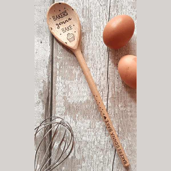 A close-up of a wooden spoon with the phrase "Bakers gonna bake" engraved on the handle. The spoon is well-worn, showcasing its years of use in the kitchen. Its smooth surface and rounded edges indicate its durability and comfortable grip. The natural color and grain of the wood add a rustic charm to the spoon, complementing the playful and whimsical message.