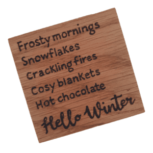 A square wooden coaster featuring intricate oak woodwork with the words 'Hello Winter' engraved on the surface in elegant script. The coaster is handcrafted with attention to detail, showcasing the warmth and natural beauty of wood grain