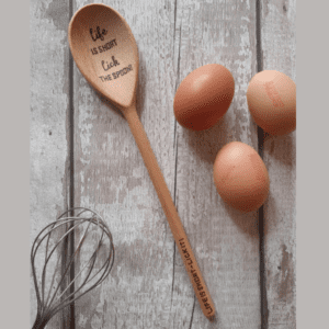 Close-up of a wooden spoon with a playful message engraved on it. The spoon reads, 'Life is short, lick the spoon.'