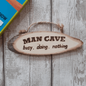 "A wooden hanging plaque for a man cave, featuring a rugged, rustic design. The plaque is oval in shape with rustic edges, giving it a vintage look. It is made of high-quality wood and features hand burnt lettering that reads 'Man Cave' in a masculine font. The background of the plaque is textured with a weathered finish, adding to its rustic charm. A sturdy rope is attached to the top corners, allowing for easy hanging on a wall or door. The perfect addition to any man cave decor, showcasing a sense of ruggedness and personal space."