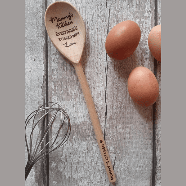 A close-up image of a wooden spoon with a smooth, polished handle and a rounded, spoon-shaped end. Engraved on the handle is the text, "Mummy's Kitchen," while the spoon end bears the words, "Everything is stirred with love." The rich, warm tones of the wood grain add a touch of rustic charm to the spoon's overall appearance.