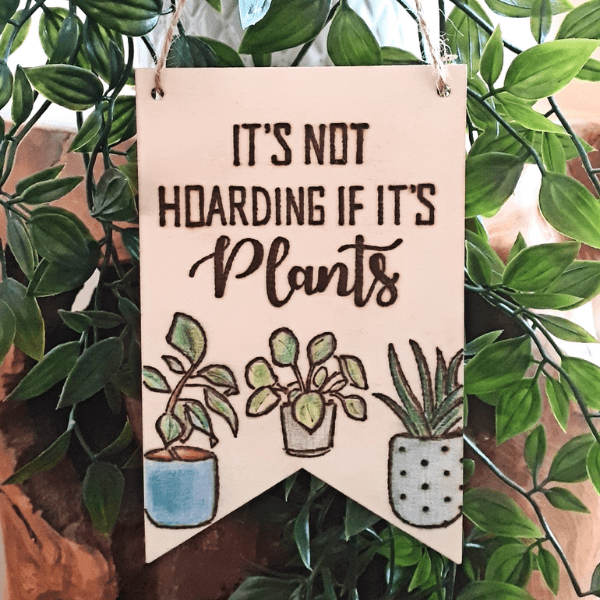 A wooden sign with black lettering that reads, "It's not hoarding if it's plants." The sign is hanging on a wall and has a plant design