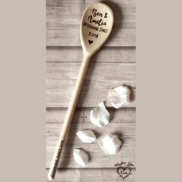 A close-up image of a wooden spoon with a hand-burnt inscription that reads "Spooning since [insert personalized names and date]". The spoon features a smooth, polished surface and showcases intricate grain patterns. The handwritten text stands out, revealing a personal touch on the otherwise plain utensil.