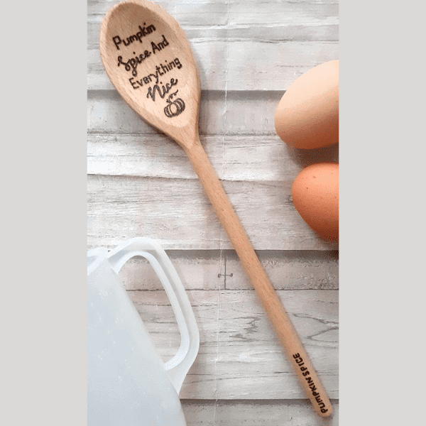 A wooden spoon resting on a rustic table, adorned with a festive autumn arrangement. Engraved on the spoon's handle are the words "Pumpkin spice and all things nice," evoking the warm and cozy spirit of autumn and Halloween.