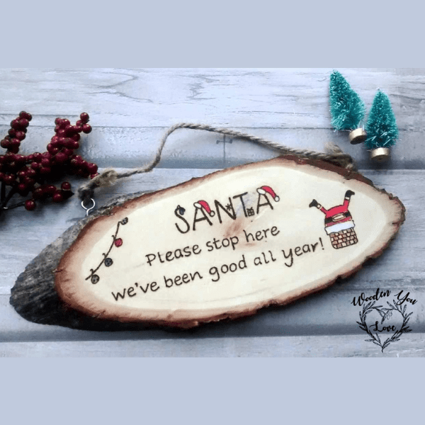 Rustic oval slice of wood with hand-burnt text that reads, "Santa please can you stop here, we have been good all year." The design features a depiction of Santa's legs popping out of a chimney.